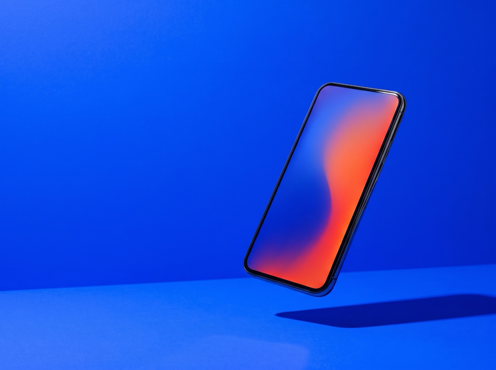 A smartphone floating at an angle above a bright cobalt blue studio backdrop, its blank screen a smooth blue-to-orange gradient, casting a hard shadow on the floor