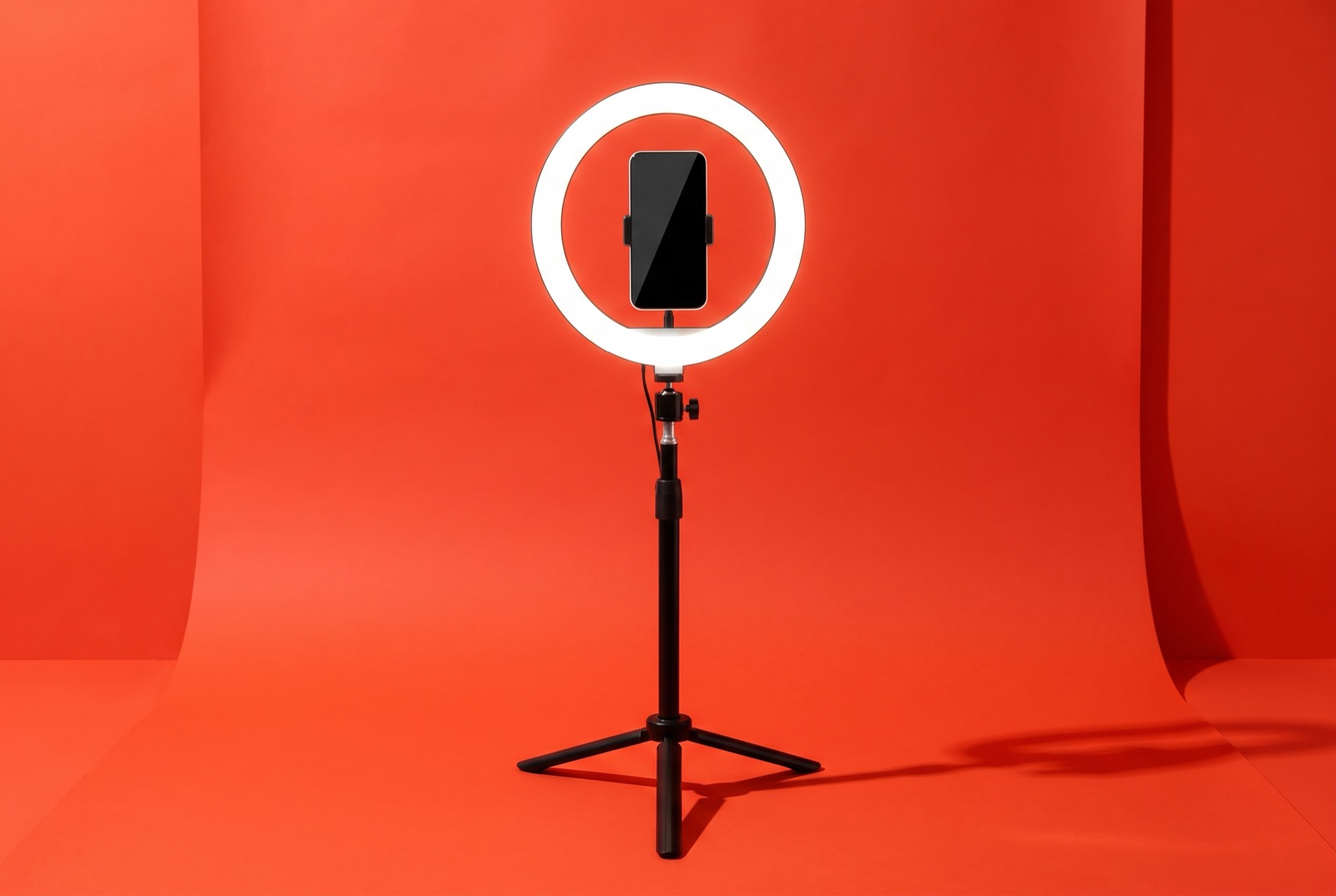 A white ring light switched on, with a smartphone clamped in its center, standing on a tripod against a bright orange studio backdrop