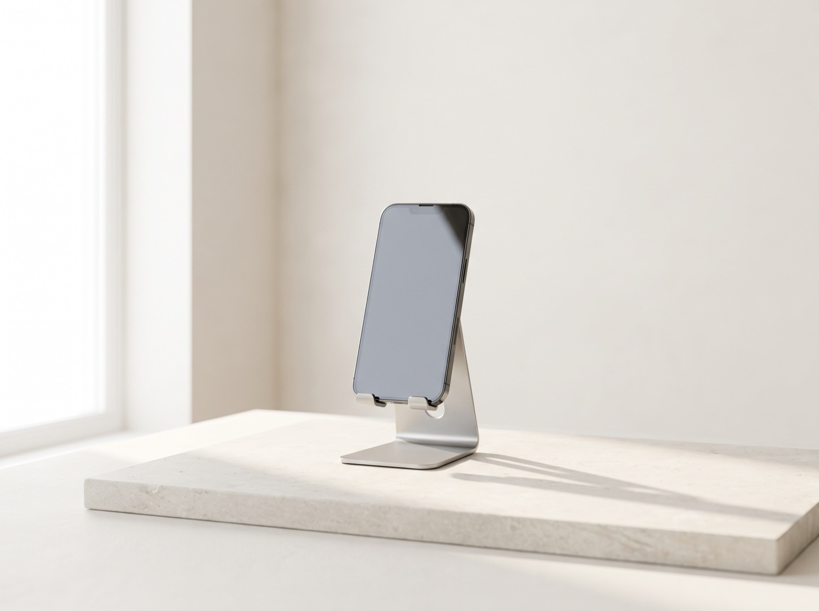 A smartphone with its screen off on a slim aluminum stand, resting on pale stone in soft morning daylight