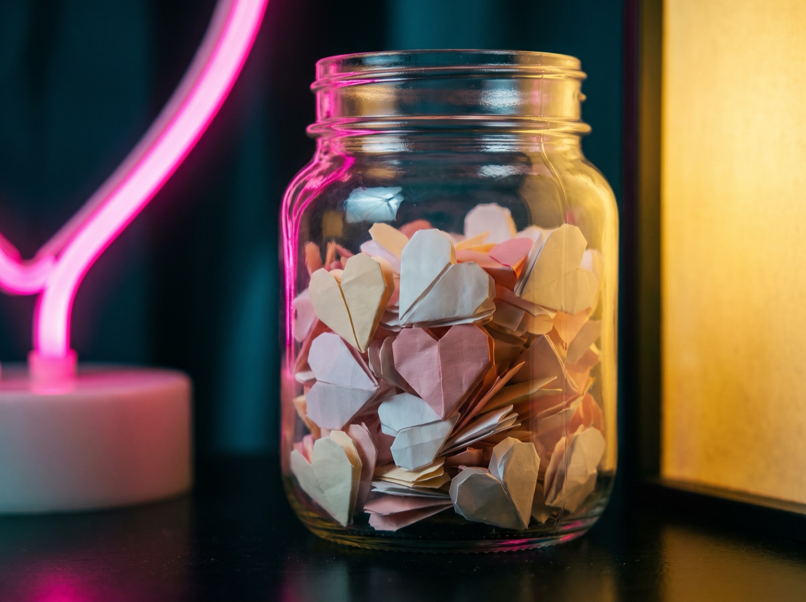 A glass tip jar of paper hearts glowing in pink and yellow neon