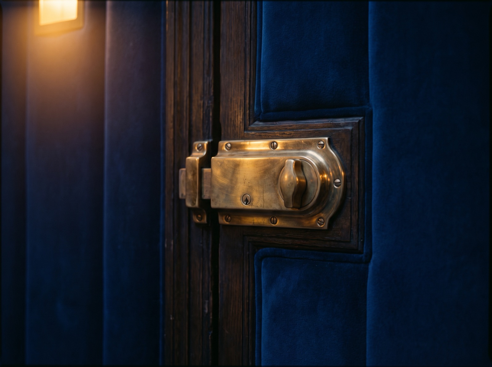 A polished brass lock on a dark wooden door lined with deep blue velvet, lit by one warm lamp