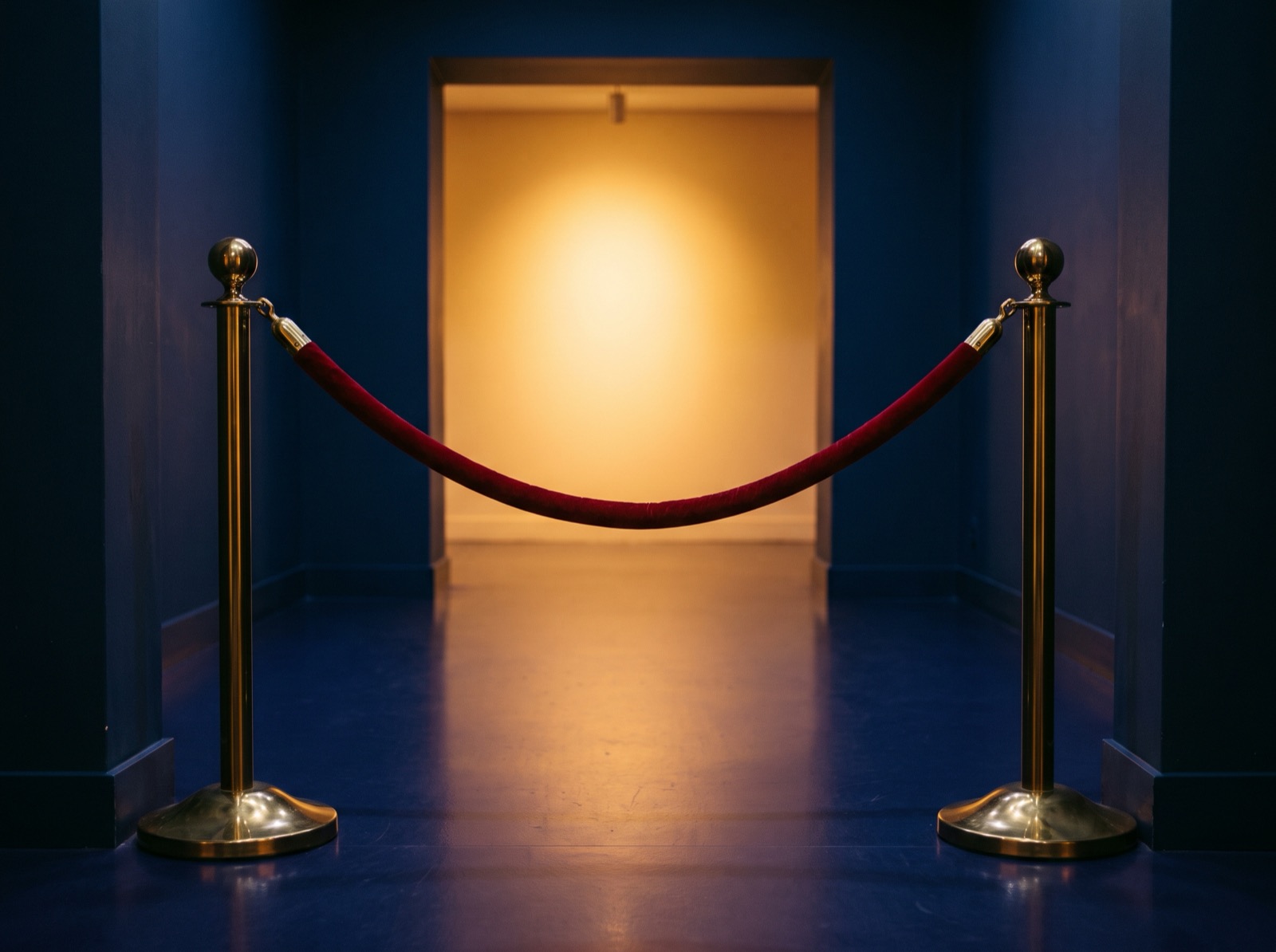 A red velvet rope on brass stands across an empty doorway, a warm amber room beyond
