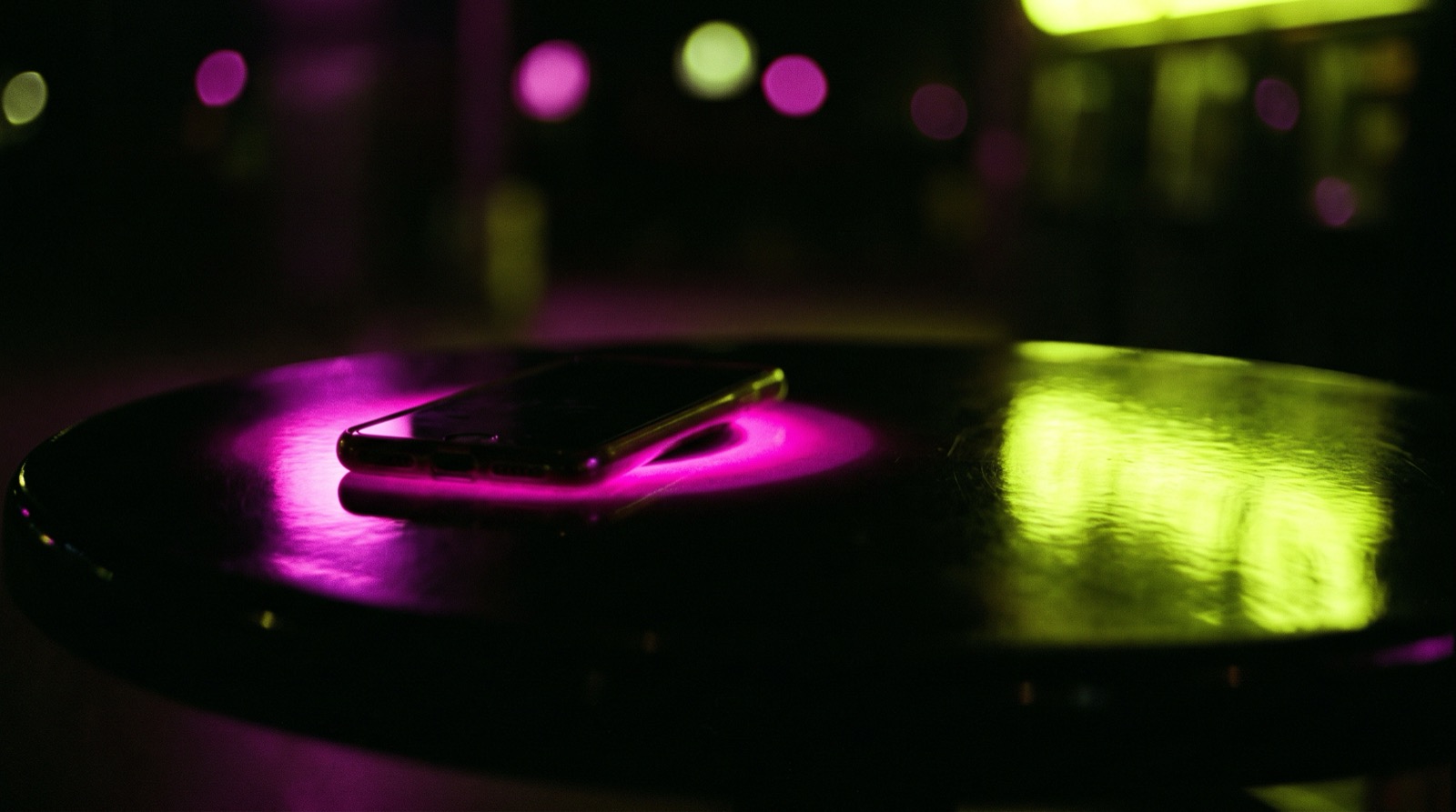 A smartphone on a glossy black table at night, hot magenta light spilling out from underneath it, lime light reflected across the tabletop